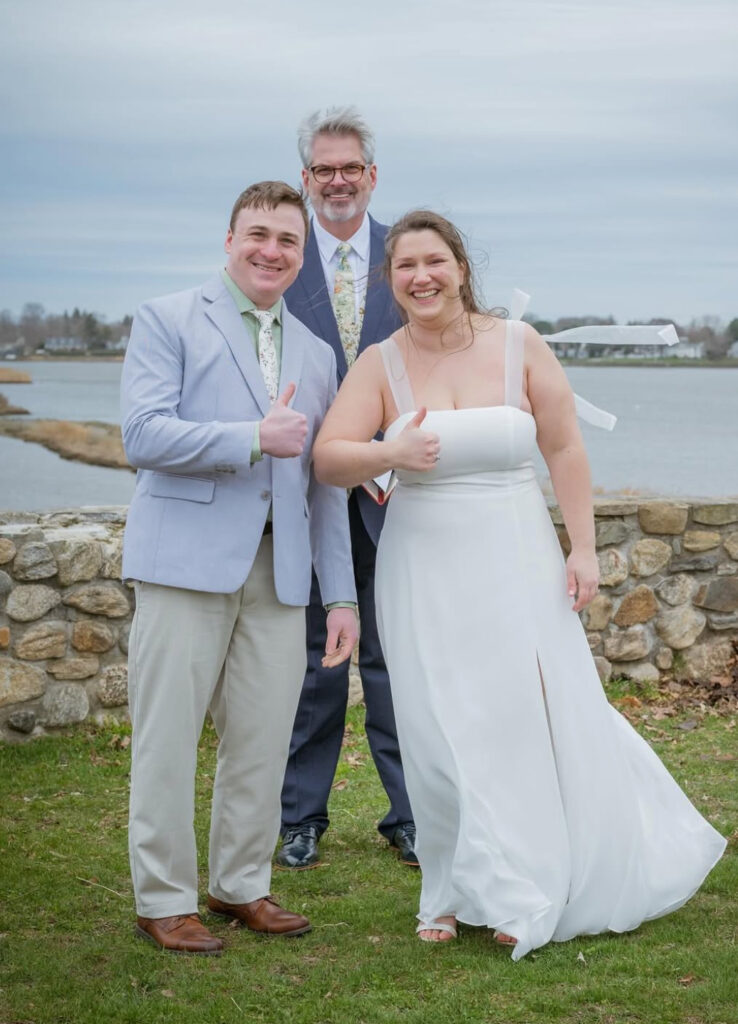 Bride in a white wedding gown poses with two men in light suits outdoors by a stone wall and lake, all smiling and giving thumbs up.