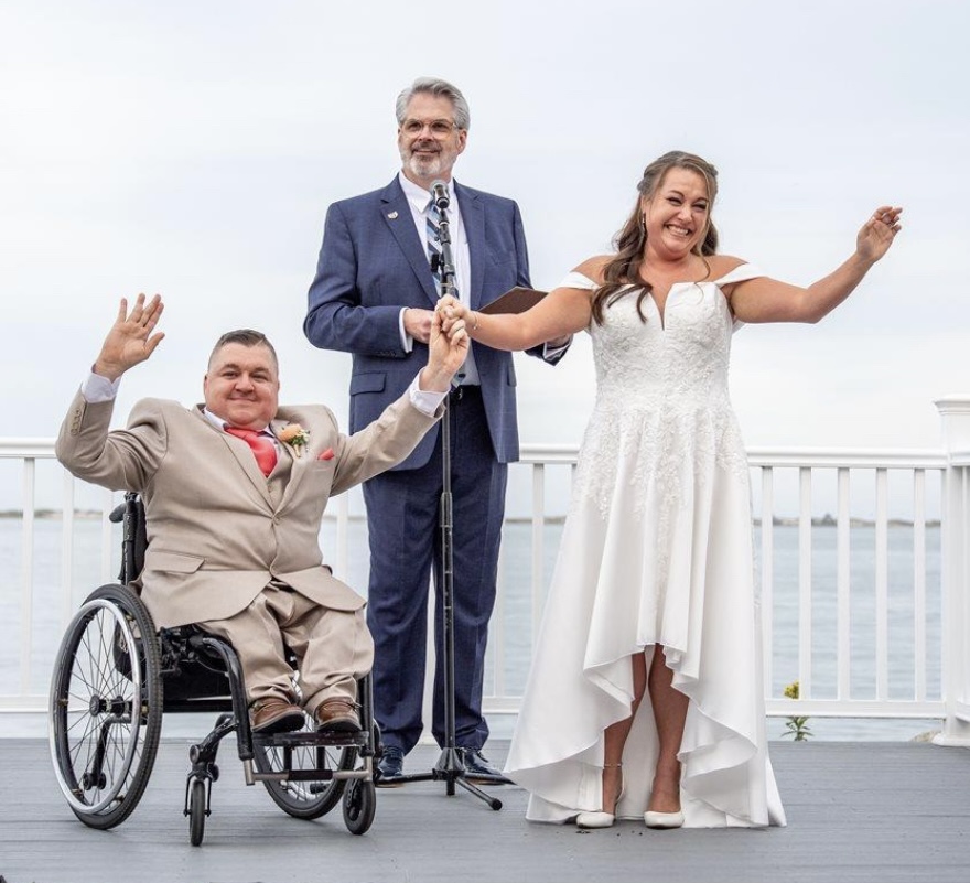 Bride in a white wedding dress and a groom in a wheelchair celebrate outdoors with an officiant, by a waterfront railing.